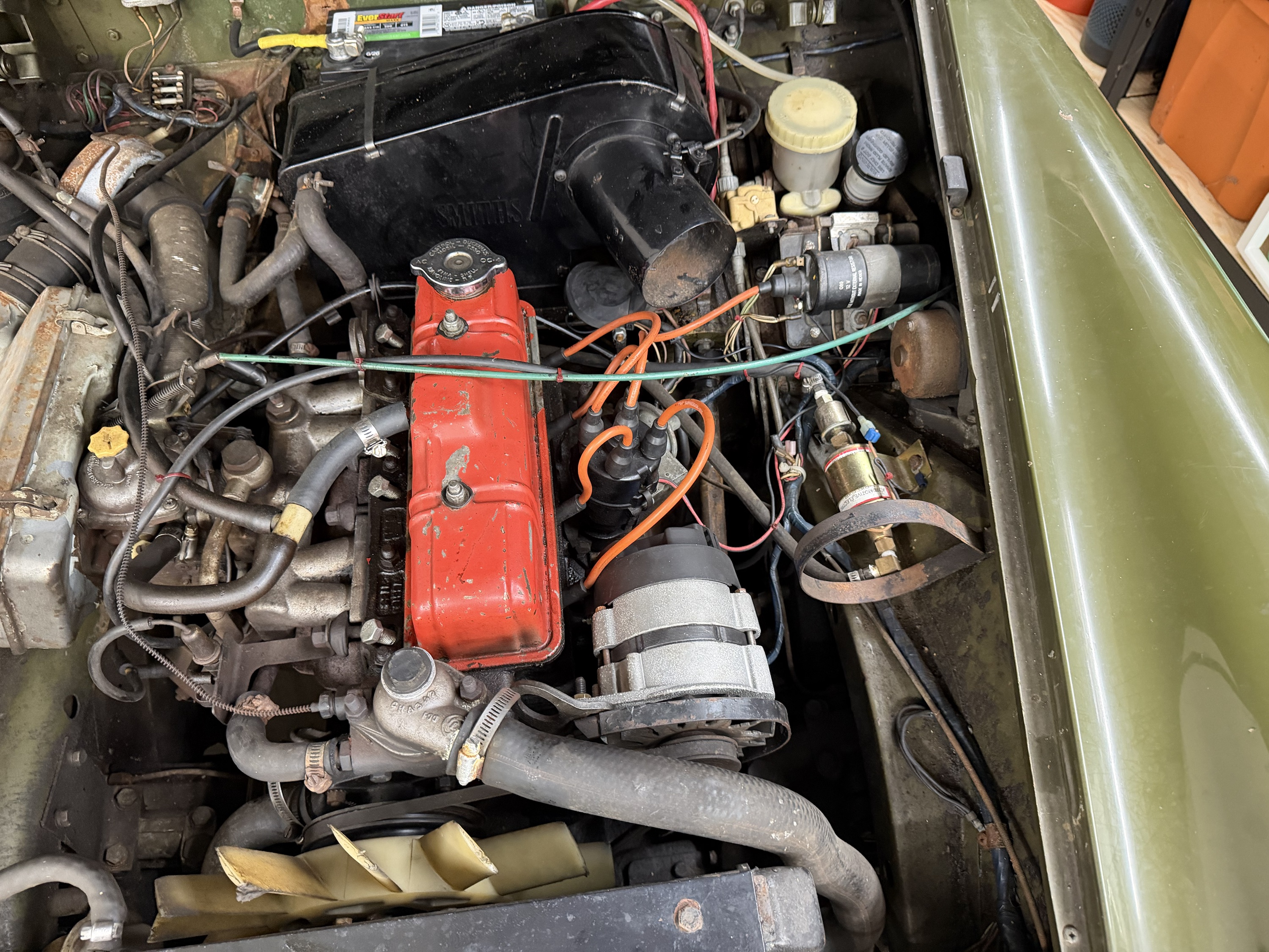 The Midget’s engine bay: a worn red rocker cover, orange ignition leads, hoses and an olive-green wing.