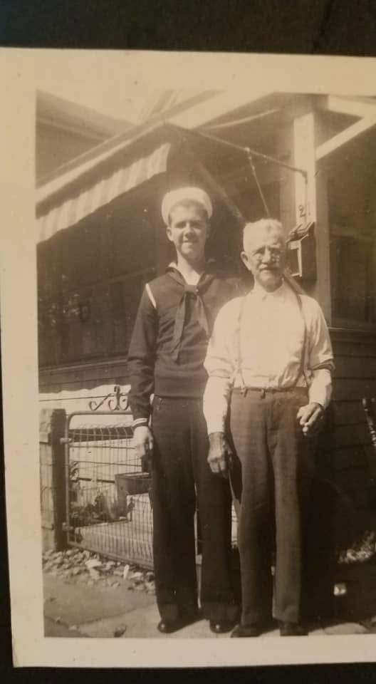 A young sailor in United States Navy dress uniform beside an elderly man in shirtsleeves and braces, on a wooden porch